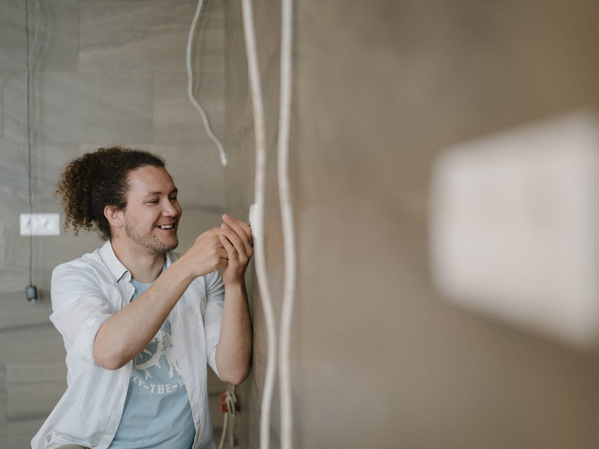 An electrician with curly hair adjusts an outlet indoors, smiling as he works.